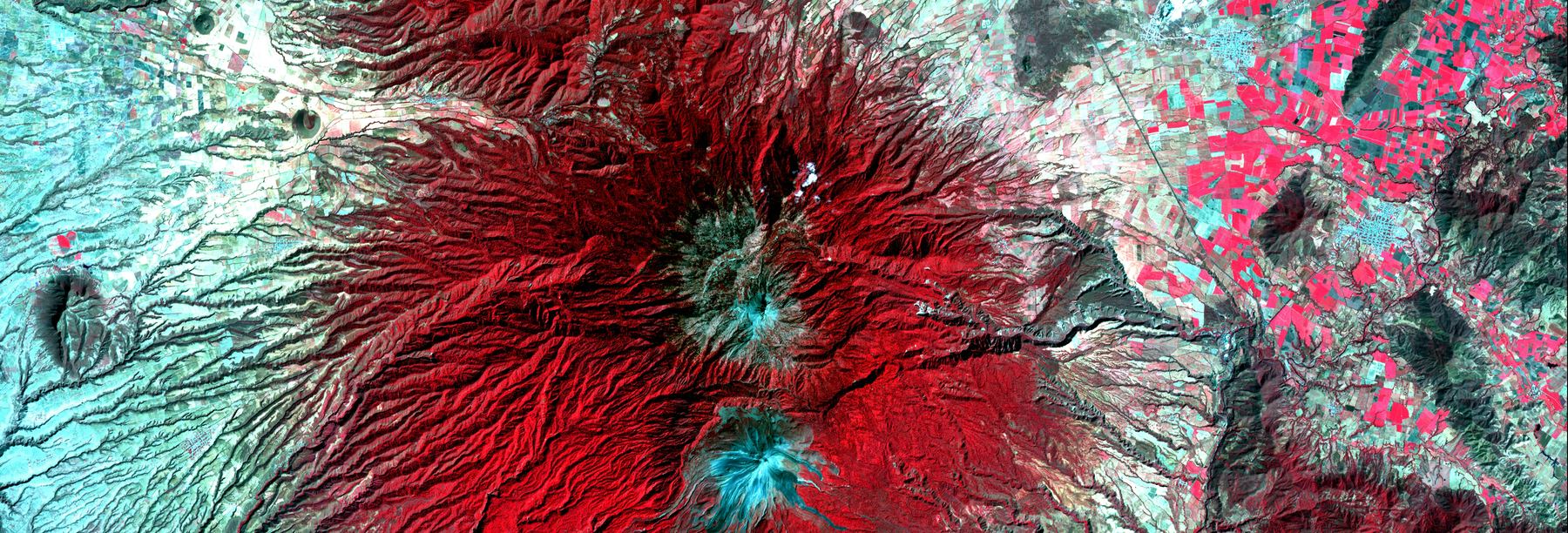 False-color satellite image of a volcanic cone in deep red, erosion channels radiating from a pale crater.